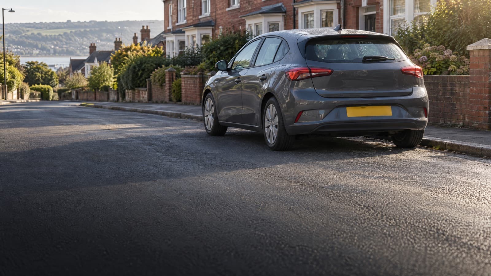 Grey hatchback parked on a quiet red-brick Devon street in low morning sun, seen from the rear with the estuary beyond